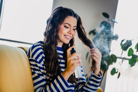 Pretty woman styling her hair with spray in a cozy living room during a relaxing weekend indoors
