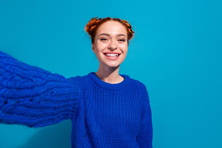 Smiling young woman taking a selfie wearing a blue knit sweater against a colorful vibrant studio backdropの写真素材