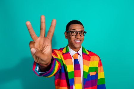 Young man in a vibrant rainbow suit smiling on a turquoise background holding up three fingersの写真素材