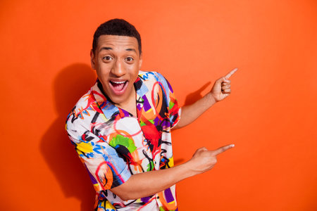 Joyful young man in vibrant printed shirt pointing with excitement against orange background, showcasing trendy summer fashionの写真素材