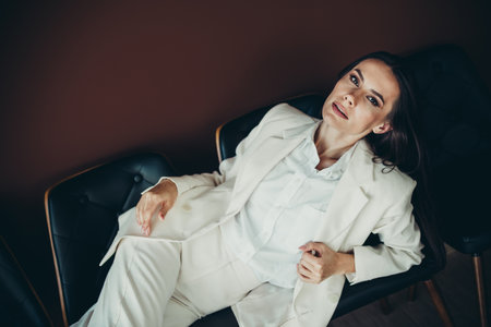 Stunning businesswoman in a white suit reclining confidently against a brown background exuding...