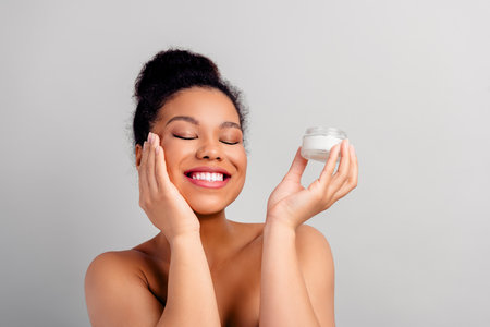 Smiling young woman with natural hairstyle holding facial cream, promoting skincare routine for radiant skinの写真素材