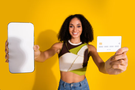Smiling woman holding smartphone and a credit card on vibrant yellow background, promoting e-commerce and digital paymentsの写真素材