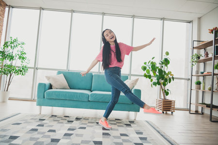 Young Asian Woman Enjoys Leisure Time Dancing Energetically in a Bright Living Room with Comfortable Home Decorの写真素材