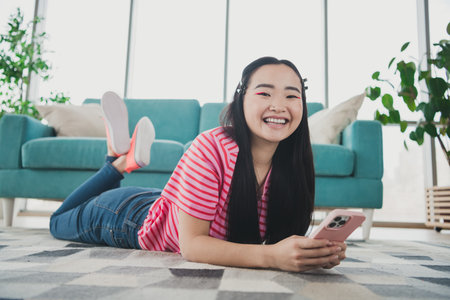 Young woman relaxing at home with her smartphone, lying on the floor of a bright living room, enjoying leisure time during a sunny dayの写真素材