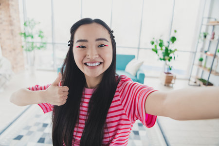 Young woman in pink striped t-shirt showing thumbs up with cheerful expression in bright living room with stylish decor, natural light.の写真素材