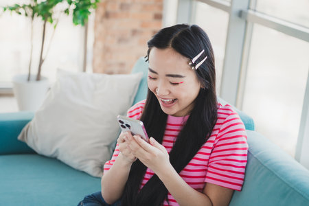 Happy young woman indoors using a smartphone while relaxing on a couch in a cozy home environment on a bright dayの写真素材