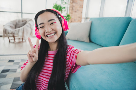 Young woman enjoying music with headphones while making a peace sign and smiling in a cozy living room interior with a turquoise sofaの写真素材