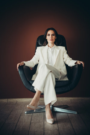 Beautiful young businesswoman in elegant white suit seated in black leather chair against brown backgroundの写真素材