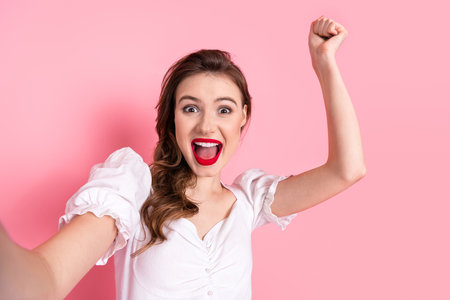 Happy young woman celebrating with raised arms against pink background wearing a stylish white dressの写真素材