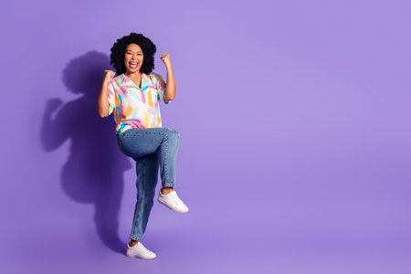 Cheerful young woman in a colorful outfit celebrating with enthusiasm against a bright purple studio backgroundの写真素材