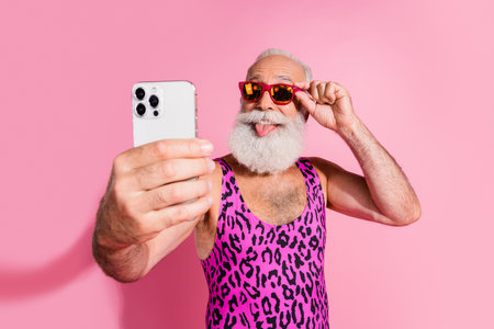 Photo of charismatic elderly cool man take selfie photo stick tongue out wear leopard print overall isolated on pink color backgroundの写真素材