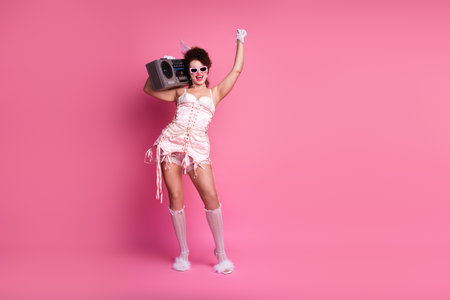 An energetic young woman embodies glamour and retro fashion in a pink background, holding a boombox.