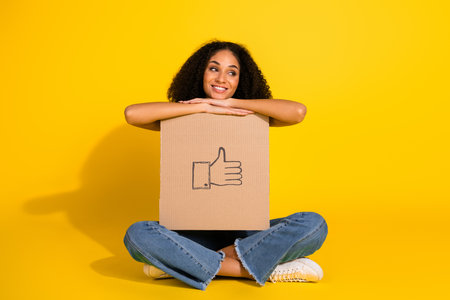 Positive young woman with curly hair holding a cardboard featuring thumbs-up symbol against vibrant...