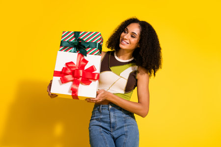 Young woman holding decorated gift boxes in front of yellow background, showcasing style, joy, and festive spiritの写真素材