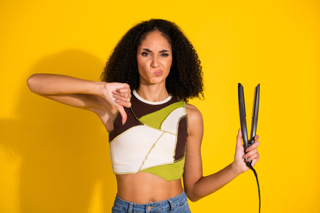 Young woman with curly hair expressing dislike for hair straightener tool on a vibrant yellow...