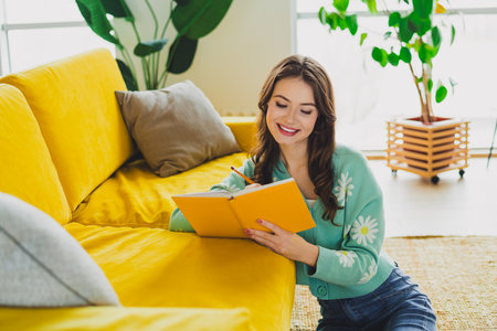 Young woman writing in a notebook while relaxing in a cozy room with natural daylight and stylish...