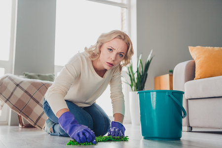 Young woman cleaning the floor at home wearing gloves and a determined expression showing effort and dedication in a living room settingの写真素材