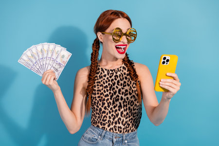 Happy young woman with red hair holding cash and phone, wearing leopard print top and sunglasses,...
