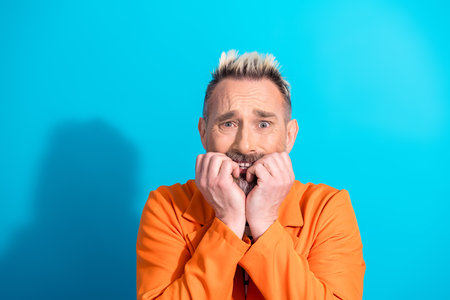 Middle aged man with dyed blond hair in an orange suit gestures anxiously on a vivid blue background...
