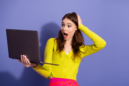 Surprised woman wearing yellow cardigan holds laptop in disbelief against purple background,...