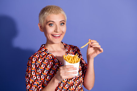 Cheerful young woman with short blond hair enjoying french fries against a vibrant purple background, wearing a colorful patterned shirtの写真素材