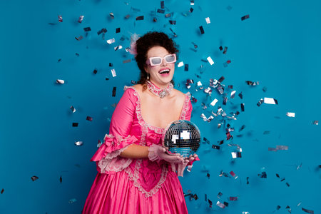 Joyful woman in an ornate pink corset with a disco ball surrounded by confetti and set against a...