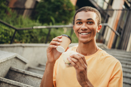 Young man enjoying a coffee and sandwich outdoors on city steps during a sunny dayの写真素材