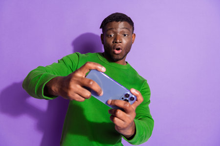 Young man in a green sweatshirt holding a smartphone excitedly against a vibrant purple background, emphasizing casual styleの写真素材