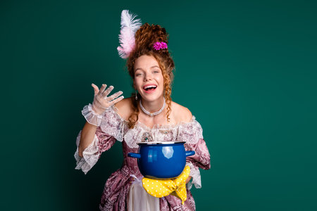 Young woman in baroque outfit displaying cheerful expression while holding a blue pot against green backgroundの写真素材