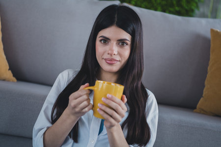 Young woman with long dark hair relaxing indoors with a yellow mugの写真素材