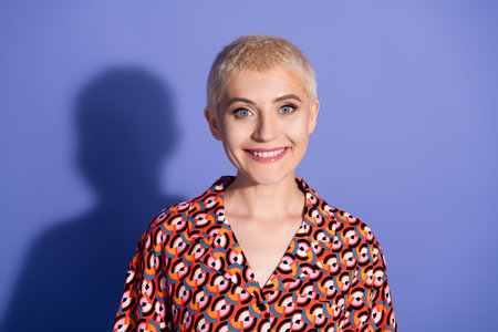 Confident young woman with short blond hair in patterned shirt smiling against a purple background,...
