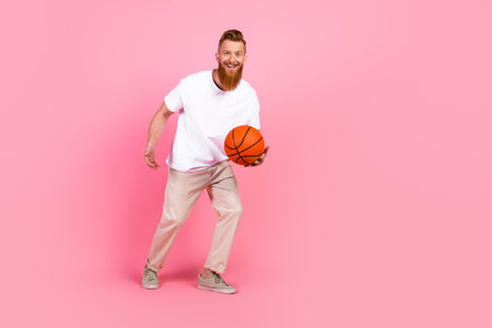 Smiling man with red beard holding a basketball on pink background in casual attire promoting an...