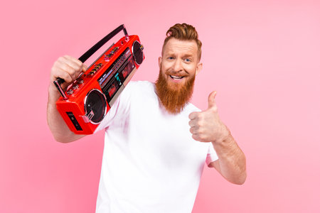 Casual young man with a red beard holding a retro red boombox and gesturing thumbs up on a pink...