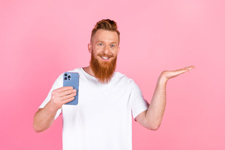 Happy young man with red beard holding smartphone and gesturing confidently against pink backgroundの写真素材
