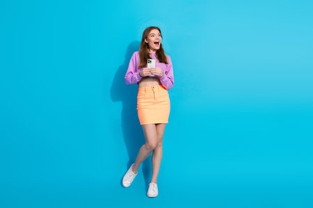 Excited young woman holding smartphone in bright modern outfit against blue background, showcasing...
