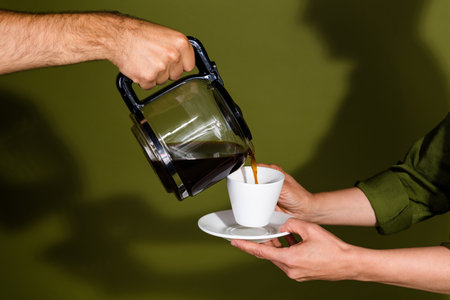 Pouring hot coffee into a white cup held on a saucer with a dark green background and hands present in the elegant serving momentの写真素材