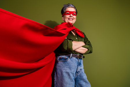 Confident senior woman in red superhero cape smiling against green wall in casual attire, showing empowerment and fun.の写真素材