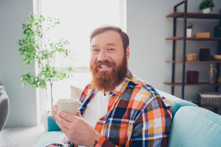 Casual bearded man enjoying coffee at home with a cheerful smile, seated in a cozy indoor living room environmentの写真素材