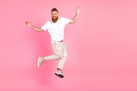 Young man with red hair and beard jumping joyfully against a bright pink background, wearing casual white t-shirt and beige trousersの写真素材