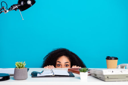 Young woman peeking nervously from behind her workspace desk, surrounded by office items against a bright solid blue backgroundの写真素材