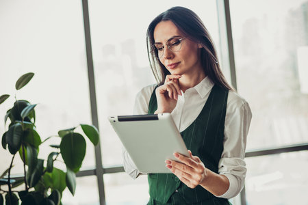 Hardworking businesswoman analyzing data on a tablet in a modern office environment with light interior and plantsの写真素材