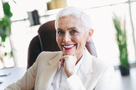Successful senior businesswoman in formal white suit seated in a modern office space, expressing elegance and professionalismの写真素材