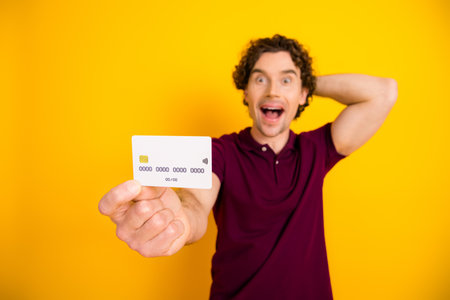 Excited young man in casual burgundy polo holding a credit card on vibrant yellow background, promotional conceptの写真素材