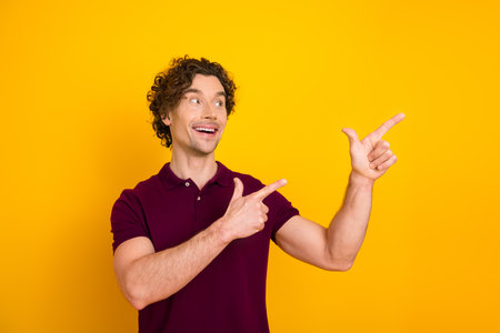 Cheerful young man with curly hair gesturing upwards on a yellow background, wearing a burgundy polo shirtの写真素材