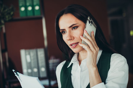Confident Businesswoman Talking on the Phone While Reviewing Documents in a Professional Office Settingの写真素材