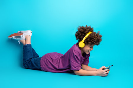 Cheerful boy with curly hair listening to music on yellow headphones while using a smartphone...