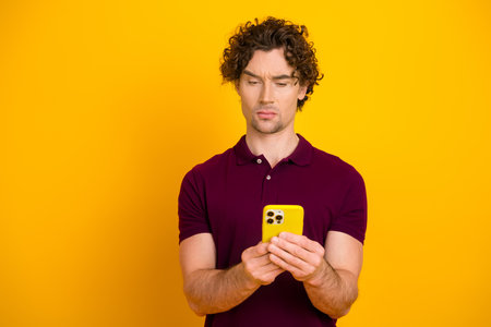 Young man with brown curly hair using a yellow phone while wearing a casual burgundy shirt on yellow...