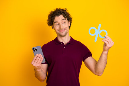 Young man holding a smartphone and a blue percentage symbol, standing against a bright yellow backgroundの写真素材
