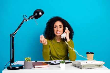 Young woman with curly hair expressing frustration while talking on a retro telephone, sitting at a desk with accessoriesの写真素材
